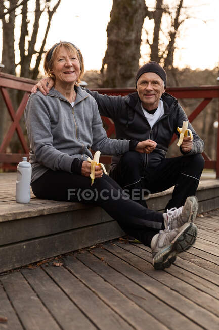 Cuerpo completo de pareja de ancianos que tienen descanso en el entrenamiento y comer alimentos saludables y mirando a la cámara - foto de stock