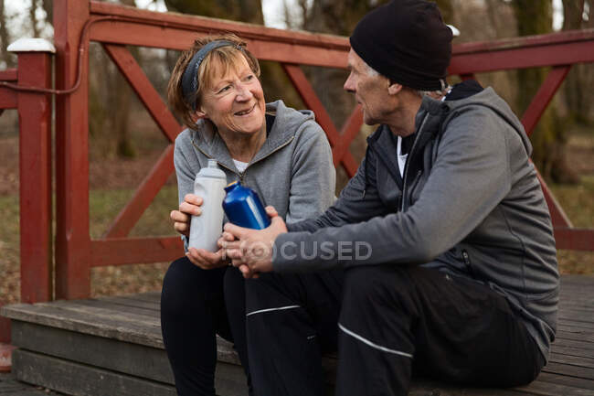Smiling senior couple sitting on wooden step with bottles of water and talking after training in park — Stock Photo