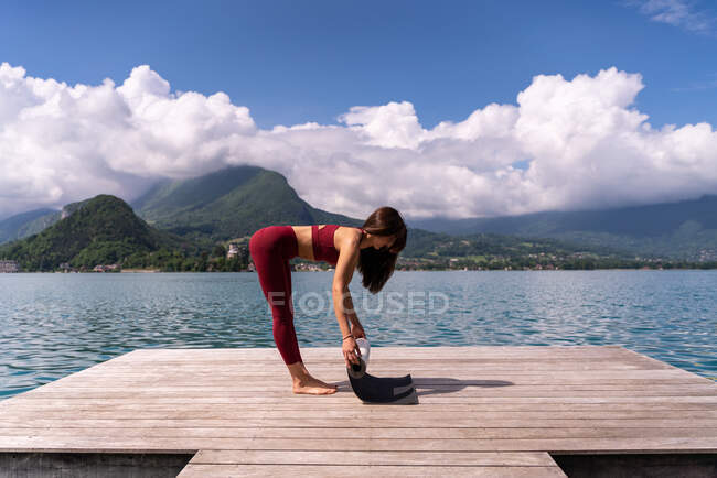 Вид збоку струнка жінка готує килимок для йоги на дерев'яній набережній біля озера влітку — стокове фото
