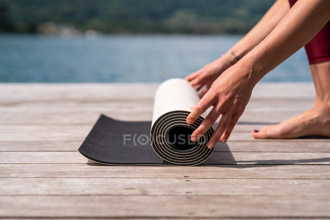 Side view of cropped unrecognizable slender female preparing mat for doing yoga on wooden quay near lake in summer — Stock Photo