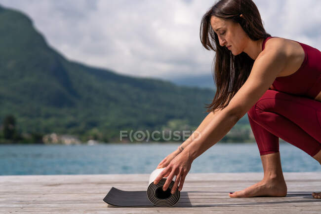 Вид збоку струнка жінка готує килимок для йоги на дерев'яній набережній біля озера влітку — стокове фото
