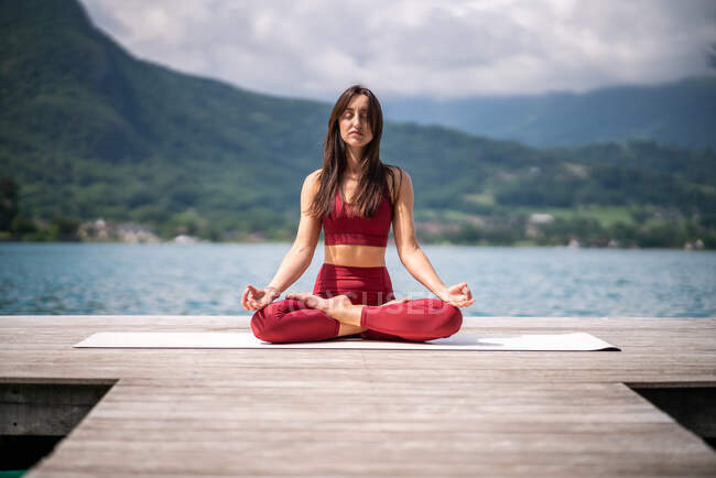 Serena fêmea sentada em Padmasana no cais de madeira e meditando com os olhos fechados enquanto pratica ioga perto do lago no verão — Fotografia de Stock