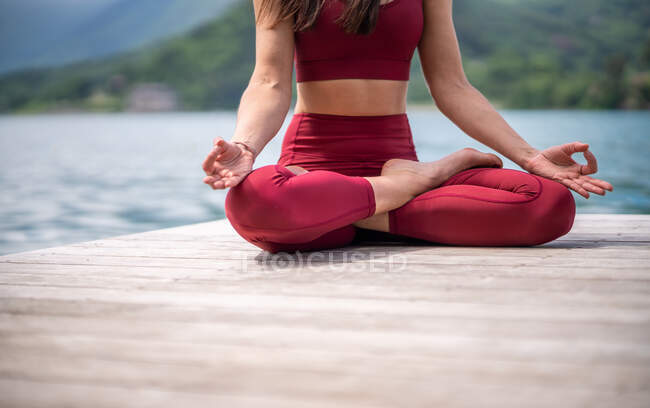 Серійна жінка сидить в Падмасані на дерев'яному пірсі і медитує під час практики йоги з жестами мудри біля озера влітку. — стокове фото