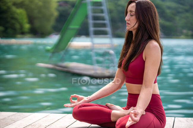 Спокойная женщина, сидящая в Падмасане на деревянном пирсе и медитирующая с закрытыми глазами во время занятий йогой у озера летом — стоковое фото