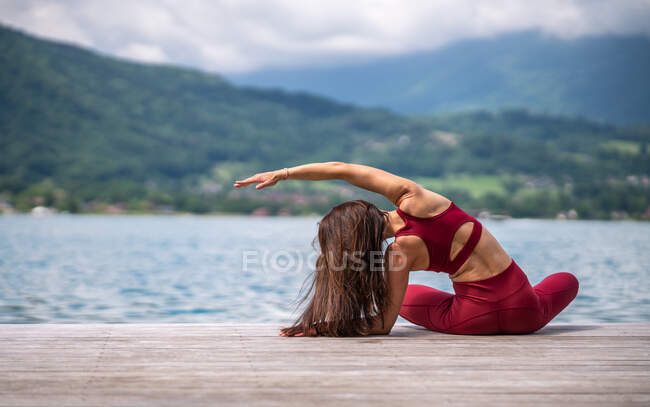 Rückenansicht einer anonymen Frau, die Yoga praktiziert und sich seitlich beugt, während sie ihren Körper streckt und am Holzkai in Teichnähe sitzt — Stockfoto