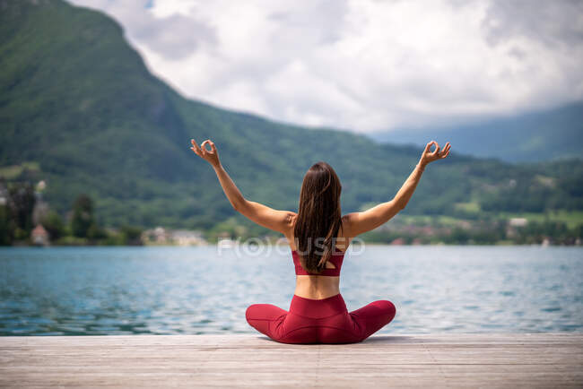 Back view of unrecognizable serene female sitting in Padmasana with arms up on wooden pier and meditating while practicing yoga with mudra gestures near lake in summer — Stock Photo