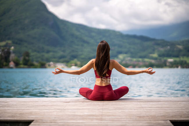 Rückansicht einer unkenntlich gelassenen Frau, die mit erhobenen Armen auf einem Holzsteg sitzt und meditiert, während sie im Sommer Yoga mit Mudra-Gesten in der Nähe des Sees praktiziert — Stockfoto