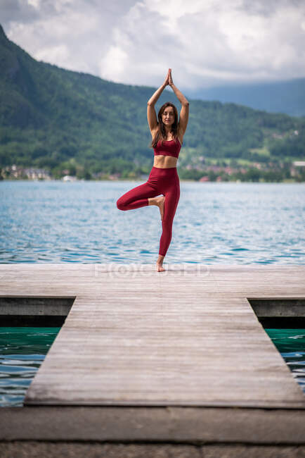 Flessibile femmina in abbigliamento sportivo praticare yoga mentre in piedi in Vrksasana con le braccia alzate sul molo vicino al lago guardando la fotocamera — Foto stock