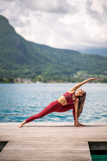 Flexible female practicing yoga in Utthita Parshvakonasana on wooden quay near lake and strecing body — Stock Photo