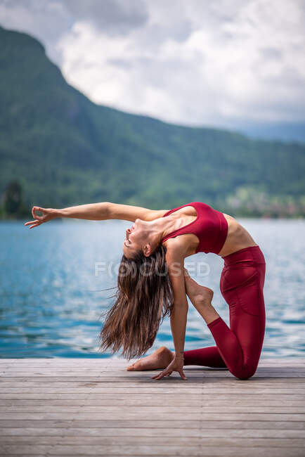 Vista lateral da pacífica fêmea praticando ioga em Ushtrasana e fazendo backbend no cais de madeira perto do lago — Fotografia de Stock