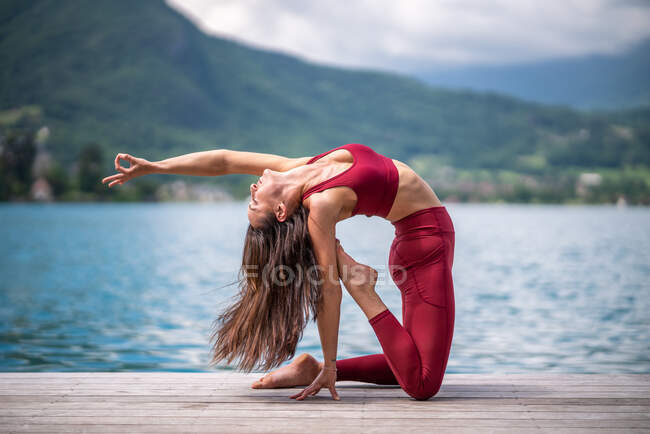 Vue latérale de paisible femme pratiquant le yoga à Ushtrasana et faisant backbend sur le quai en bois près du lac — Photo de stock