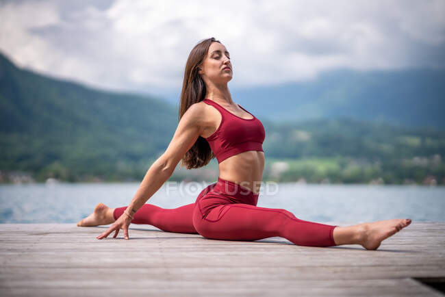 Im Erdgeschoss sitzt eine ruhige Frau in Hanumanasana auf einem hölzernen Pier, während sie im Sommer Yoga praktiziert und die Beine am See streckt — Stockfoto
