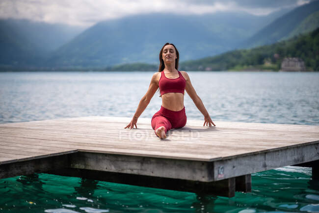 Спокойная женщина сидит в Хануманасане на деревянном пирсе, практикуя йогу и растягивая ноги у озера летом — стоковое фото