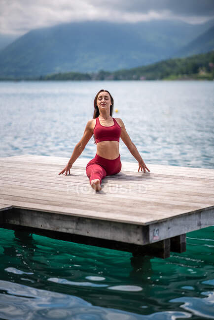 Затишна самиця сидить в Хануманасані на дерев'яному пірсі під час практики йоги і розтягування ніг біля озера влітку. — стокове фото