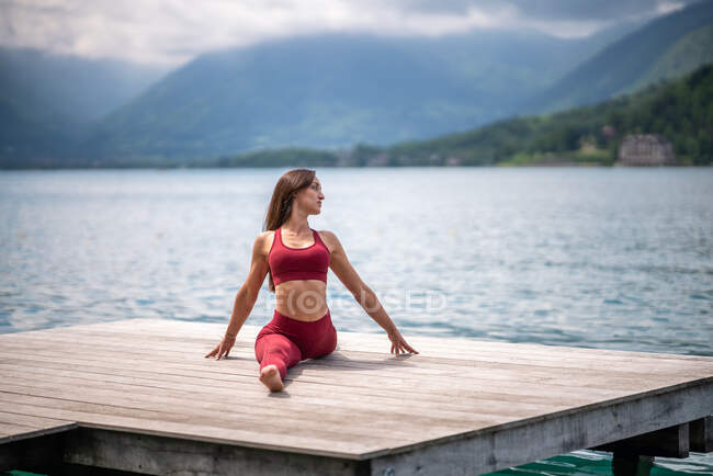 Tranquila hembra sentada en Hanumanasana en muelle de madera mientras practica yoga y estira las piernas cerca del lago en verano - foto de stock