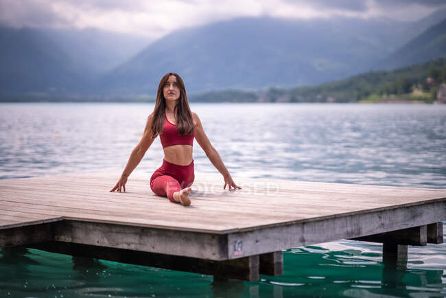 Tranquillo femminile seduto a Hanumanasana sul molo di legno durante la pratica di yoga e stretching gambe vicino al lago in estate — Foto stock