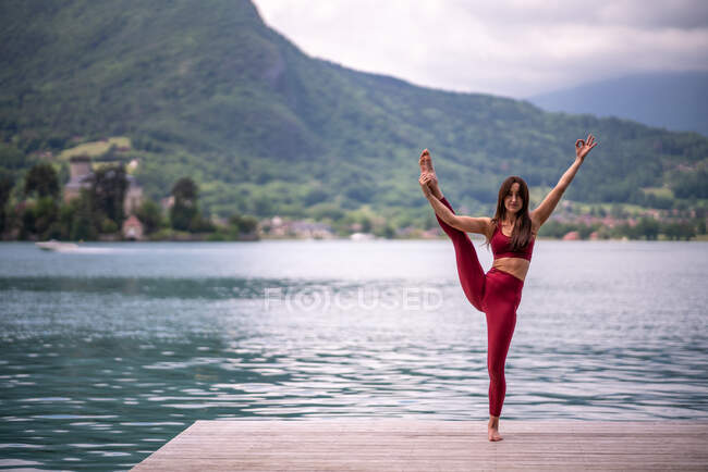 Мирная женщина балансирует на ногах в Тривикрамасане, практикуя йогу на деревянном пирсе возле пруда, глядя в камеру — стоковое фото