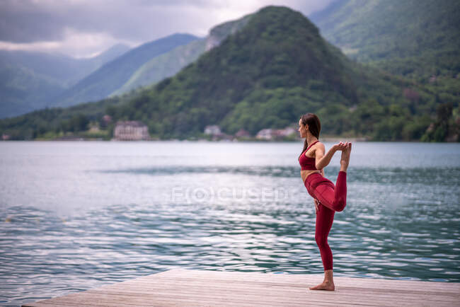 Vista lateral de pacífica hembra balanceándose en pierna en Natarajasana mientras practica yoga en muelle de madera cerca de estanque mirando hacia otro lado - foto de stock