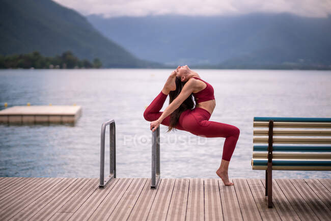 Side view of serene female in sportswear stretching legs and doing backbend while practicing yoga on wooden quay near lake — Stock Photo