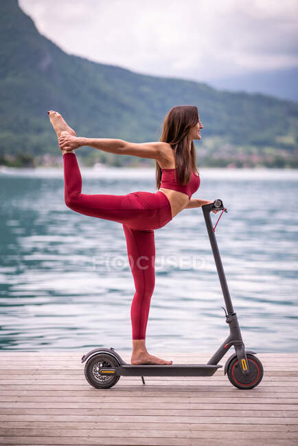 Vista lateral da fêmea flexível encantada em balanceamento activewear em Natarajasana em scooter elétrico enquanto pratica ioga no cais de madeira e olhando para longe — Fotografia de Stock
