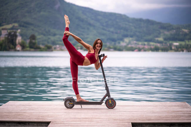 Vista lateral da fêmea flexível encantada no equilíbrio activewear em Natarajasana em scooter elétrico enquanto pratica ioga no cais de madeira e olhando para a câmera — Fotografia de Stock