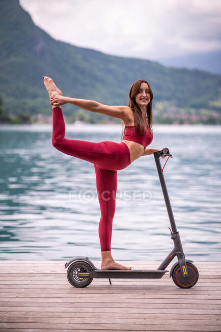 Seitenansicht der entzückten flexiblen Frau in Activwear balanciert in Natarajasana auf einem Elektroroller, während sie Yoga auf einem Holzsteg praktiziert und in die Kamera blickt — Stockfoto