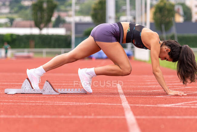 Полный вид сбоку тела сфокусированной молодой женщины-спринтера в низком стартовом положении, готовой бежать от стартовых блоков на красной дорожке стадиона — стоковое фото