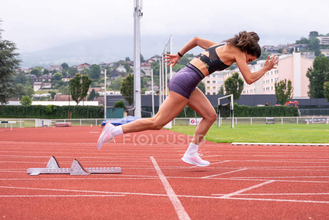 Vue latérale complète du corps d'une jeune sprinteuse déterminée qui commence à courir à partir de blocs sur la piste du stade — Photo de stock