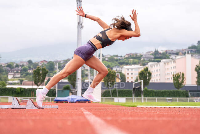 Full body side view of determined young female sprinter starting to run from blocks on track of stadium — Stock Photo
