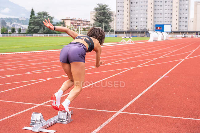 Повний вид на тіло визначеної молодої жінки-спринтера, яка починає бігати з блоків на трасі стадіону — стокове фото