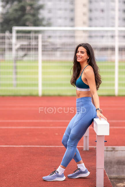 Joven atleta hispana positiva en ropa deportiva sentada en la barrera y sonriendo mientras descansa durante el entrenamiento en el hipódromo del estadio - foto de stock