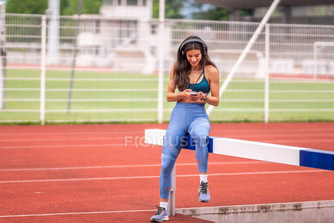 Cheerful young athletic Hispanic female in sportswear and headphones listening to music and browsing mobile phone while having break after training on stadium track — Stock Photo