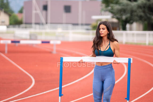 Joven atleta hispana llevando obstáculo mientras se prepara para correr con obstáculos en pista de estadio deportivo - foto de stock