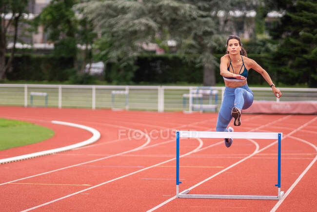 Full length of confident young Hispanic sportswoman jumping over barrier while running on red track of sports stadium — Stock Photo