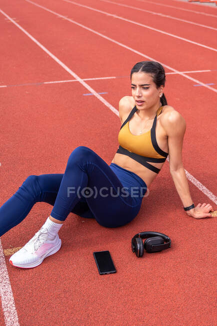 Angle élevé de jeune femme fatiguée sportive hispanique en vêtements de sport et baskets assis sur la piste rouge près du smartphone et des écouteurs tout en se reposant après l'entraînement au stade — Photo de stock