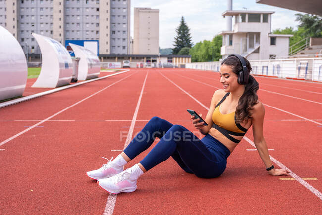 Hohe Winkel der müde junge fit hispanische Sportlerin in Aktivkleidung und Turnschuhen sitzt auf roter Bahn in der Nähe von Smartphone und Kopfhörer, während sie sich nach dem Training im Stadion ausruht — Stockfoto