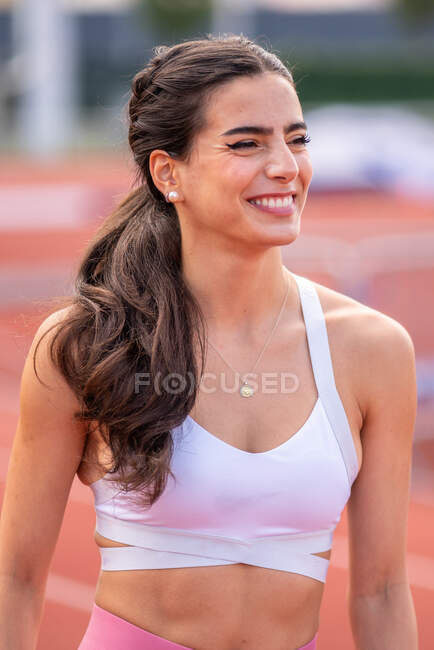Positive young Hispanic female athlete in sports top smiling and looking away while standing on blurred stadium track — Stock Photo