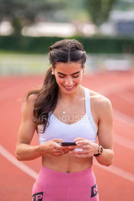 Fröhliche junge athletische Spanierin in Sportbekleidung surft während einer Trainingspause auf der Stadionbahn im Handy — Stockfoto