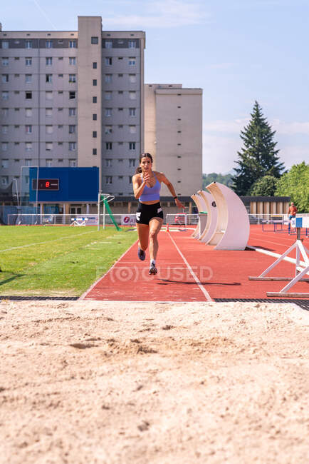 Full body of determined young female track and field athlete running fast before long jump during training on sports field — Stock Photo