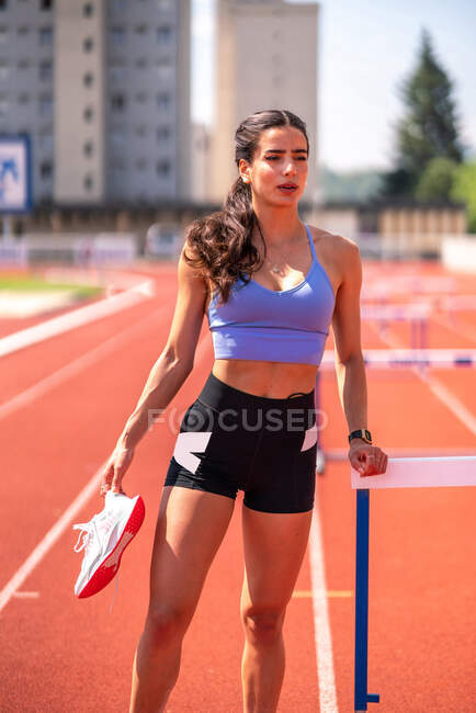 Front view of young female athlete in stylish sportswear with sneakers in hand standing near hurdle and looking away while resting after workout at stadium — Stock Photo