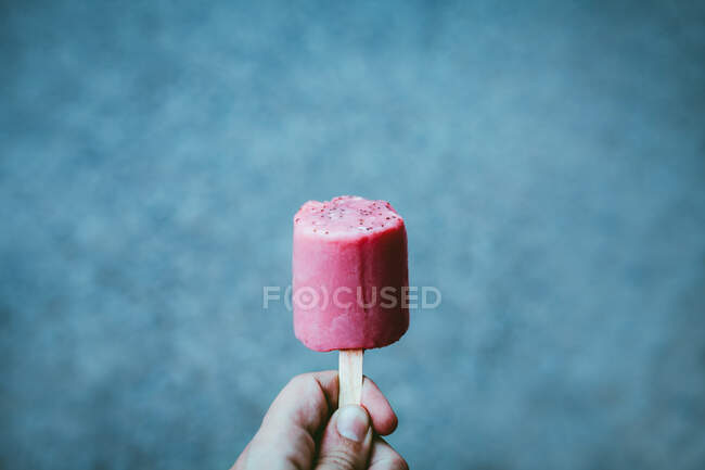 Crop Person zeigt leckere Beere Eis Pop von rosa Farbe auf verschwommenem blauem Hintergrund — Stockfoto
