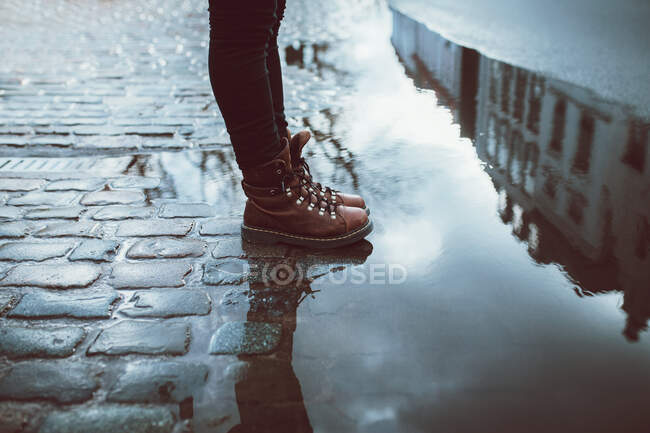 Side view of crop anonymous person in leather footwear standing on tiled walkway with puddle reflecting urban building — Stock Photo