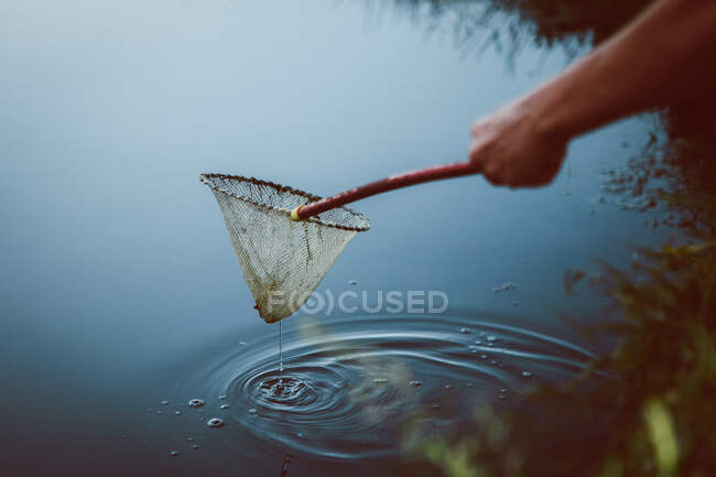 Crop unrecognizable person with net on stick catching fish in rippled lake in daytime — Stock Photo