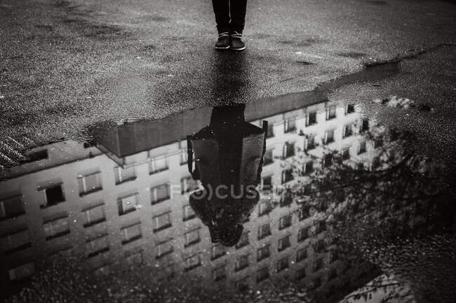 Black and white high angle of reflection of crop unrecognizable person and multistory building in puddle on wet asphalt in overcast day — Stock Photo