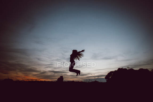 Низький кут зору на бік повний силует тіла невпізнаваної жінки з витягнутими руками та літаючим волоссям, що стрибає над землею під час заходу сонця — стокове фото