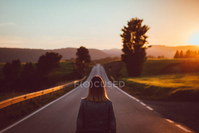 Back view of unrecognizable female standing on asphalt roadway going through green valley at sunset — Stock Photo