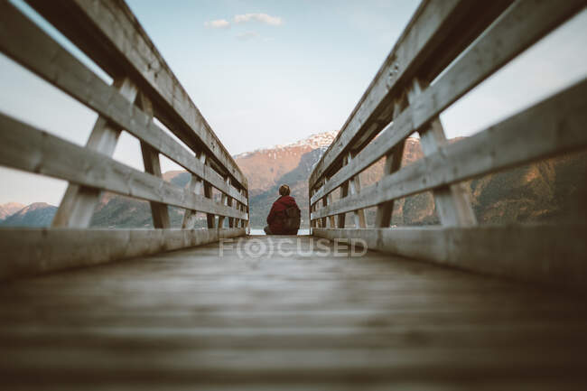 Ground level back view of unrecognizable explorer with backpack sitting on wooden pathway and admiring view of lake surrounded by mountains — Stock Photo