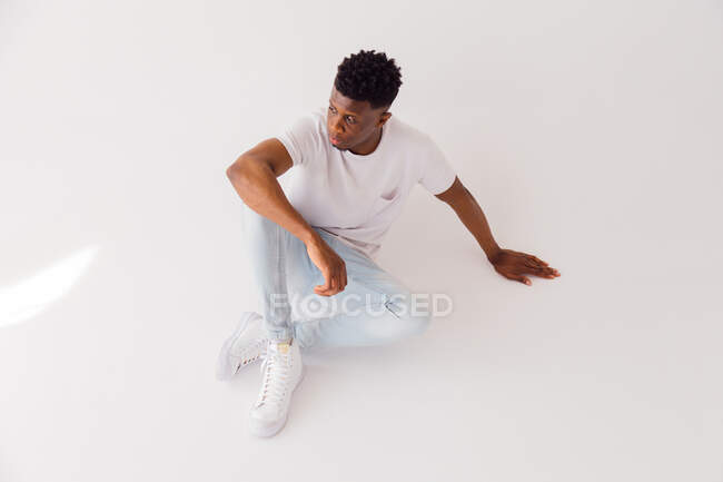 De cima corpo inteiro de jovem afro-americano modelo masculino vestindo t-shirt branca com jeans azul claro e tênis sentado no chão em estúdio — Fotografia de Stock
