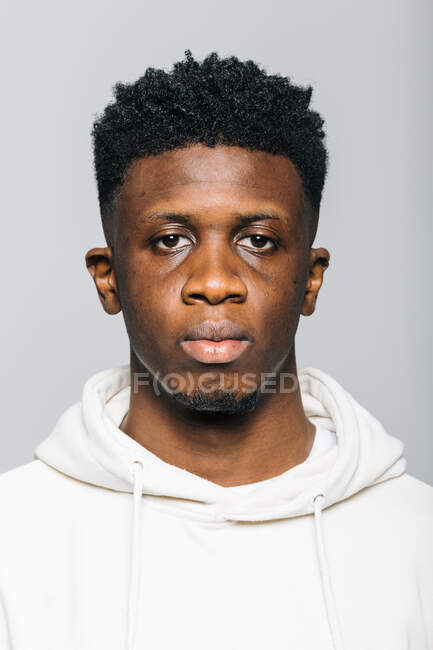 Confiant sérieux jeune homme afro-américain en sweat à capuche blanc regardant la caméra sur fond gris — Photo de stock