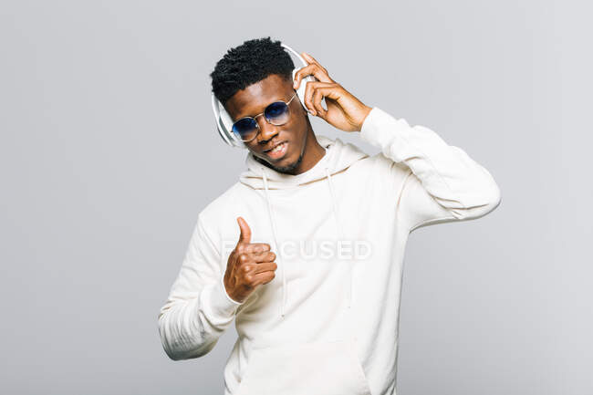 Happy young African American hipster guy in white hoodie and trendy sunglasses showing thumb up while listening to music through wireless headphones — Stock Photo